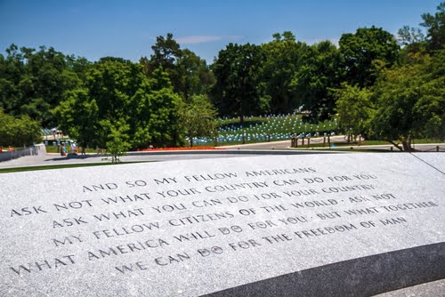 A stone engraving of JFK's famous "Ask not..." speech with a clear, blue sky and green trees in the background.