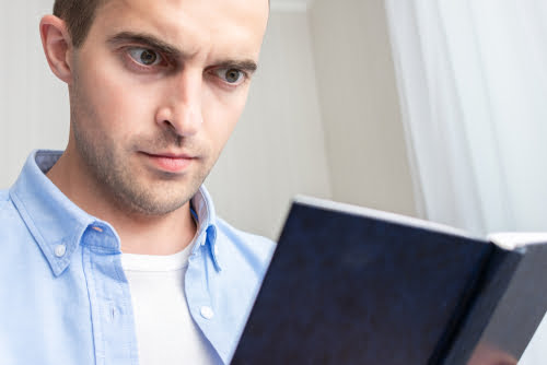 A man in a blue dress shirt reads a book and has a serious expression