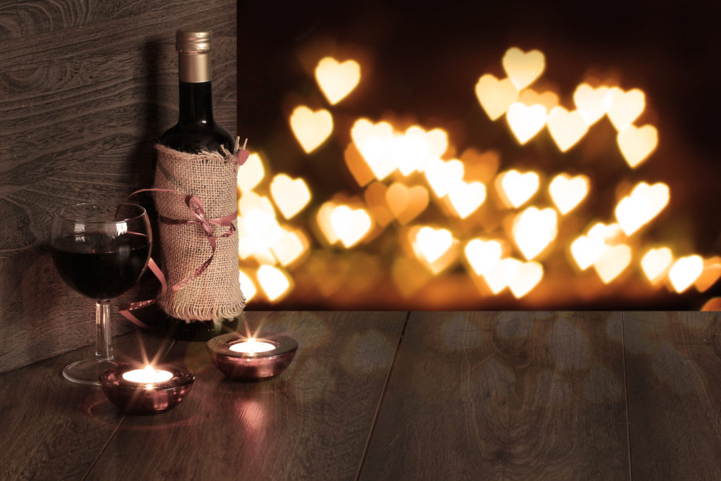 A bottle and glass of red wine paired with two candles in the foreground against soft focus, heart-shaped candle flames