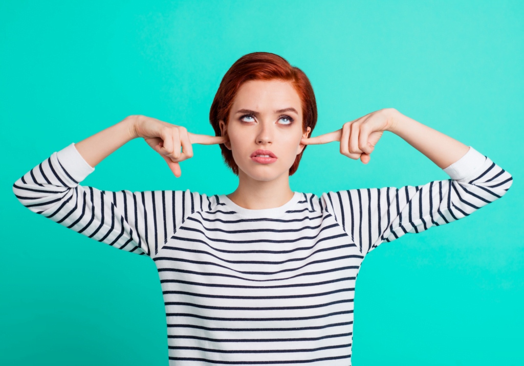 A young redhead with a pixie cut wears a black and white striped shirt and stands against a blue-green background with her index finders in her ears looking annoyed and rolling her eyes