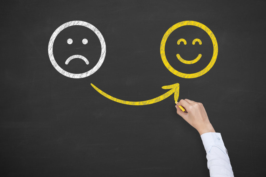 Two smiley faces on a blackboard. One is white and frowning. A hand draws a yellow arrow to another that is yellow and smiling