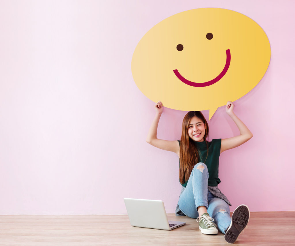 A young brunette girl wearing a dark green sleeveless t-shirt, ripped jeans, and grey sneakers sits on a hardwood floor with her laptop to one side and a giant smiley face thought bubble above her head.